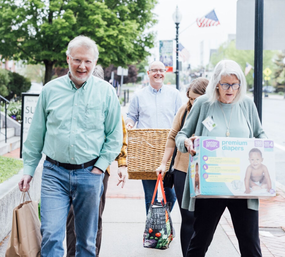 Four people walking down a sidewalk carrying containers with food and diapers.
